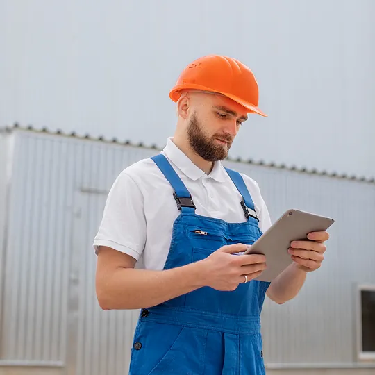 Maintenance technician in a hard hat checking a tablet outside a building