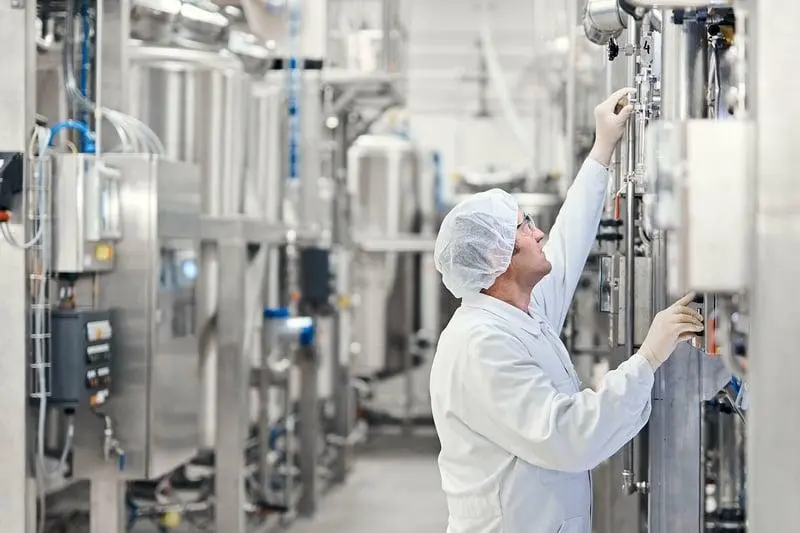 Technician in a hairnet and lab coat inspecting instrumentation on production line equipment during a scheduled maintenance check