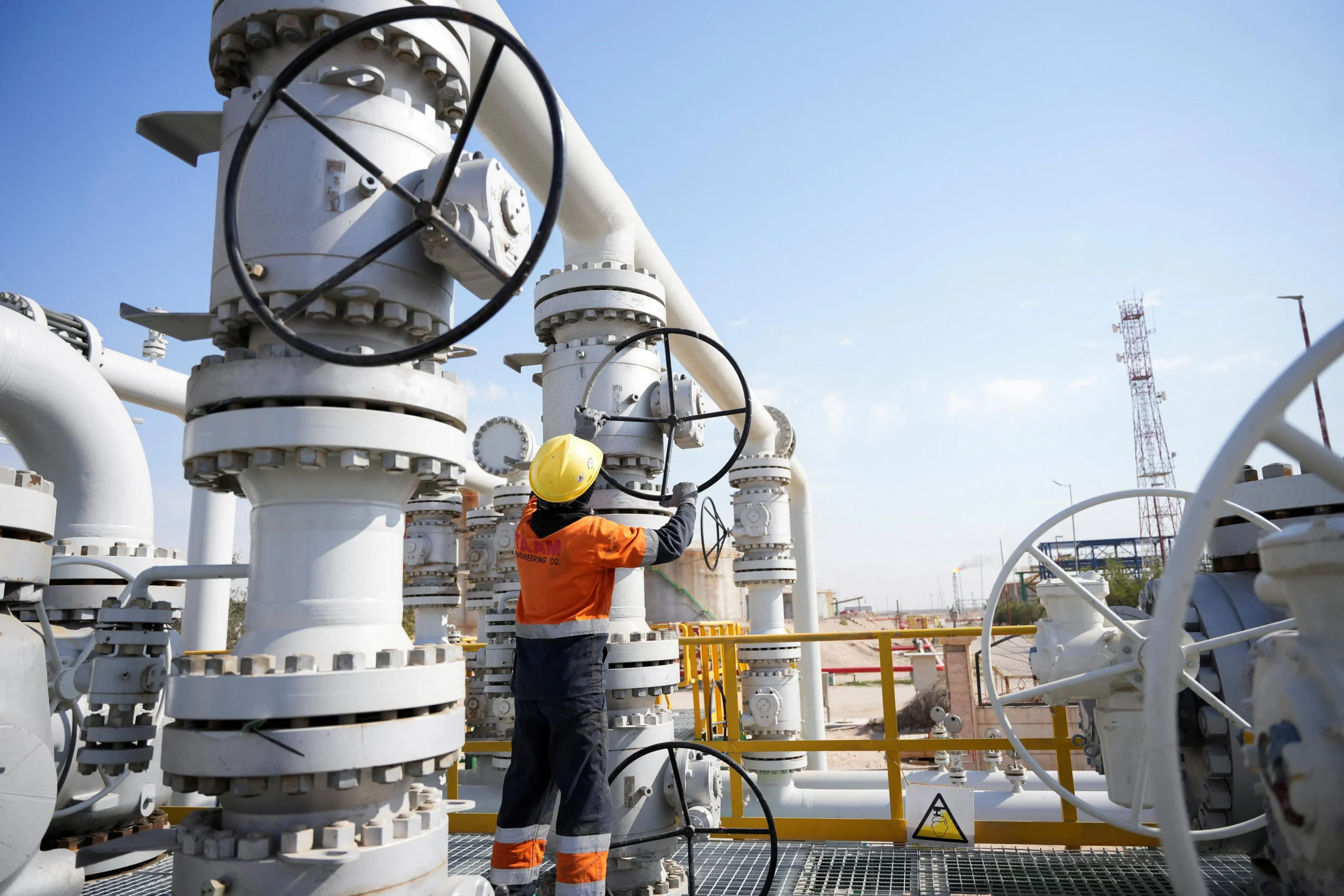 Technician in a hard hat operating a valve wheel on wellhead piping at an oil and gas processing site