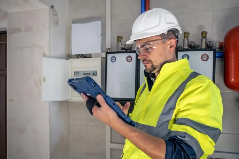 Facilities technician with a rugged tablet checking an electrical panel in a school boiler room